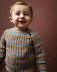 A smiling young child wearing Serendipity Organic's alpaca multicolored rib knit sweater with horizontal stripes of blue, yellow, pink, and brown over a gray base, set against a solid dark brown background.