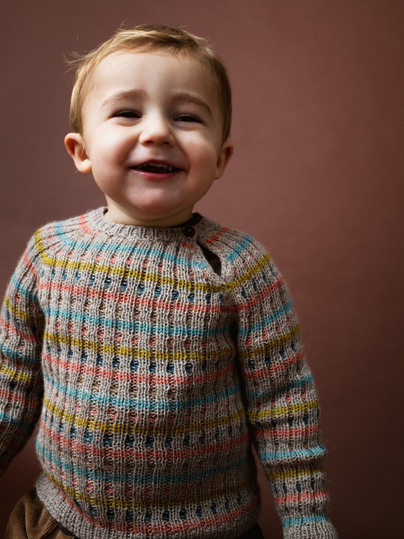 A smiling young child wearing Serendipity's alpaca multicolored rib knit sweater with horizontal stripes of blue, yellow, pink, and brown over a gray base, set against a solid dark brown background.
