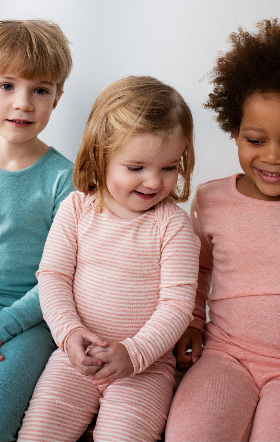 Three children wearing Serendipity organic cotton. Two on either side are wearing solids not carried by Bigelow Lane and are cropped in image. The child in the middle is wearing an organic cotton long sleeved coral stripe body suit with matching leggings