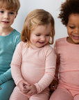 Three children wearing Serendipity organic cotton. Two on either side are wearing solids not carried by Bigelow Lane and are cropped in image. The child in the middle is wearing an organic cotton long sleeved coral stripe body suit with matching leggings