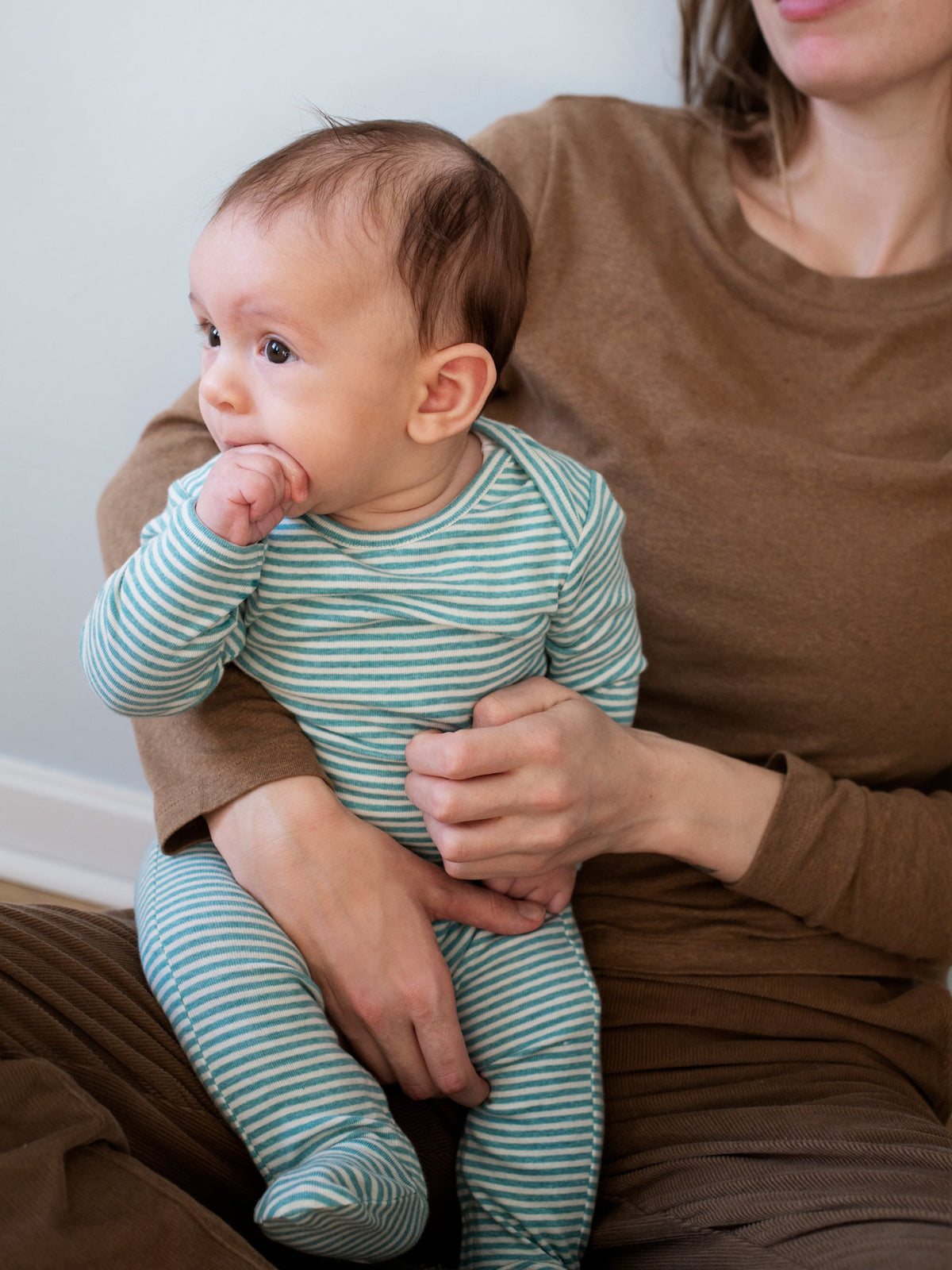 A baby wearing a serendipity organic cotton ivy striped long sleeve footed baby suit sits on an adult's lap. 