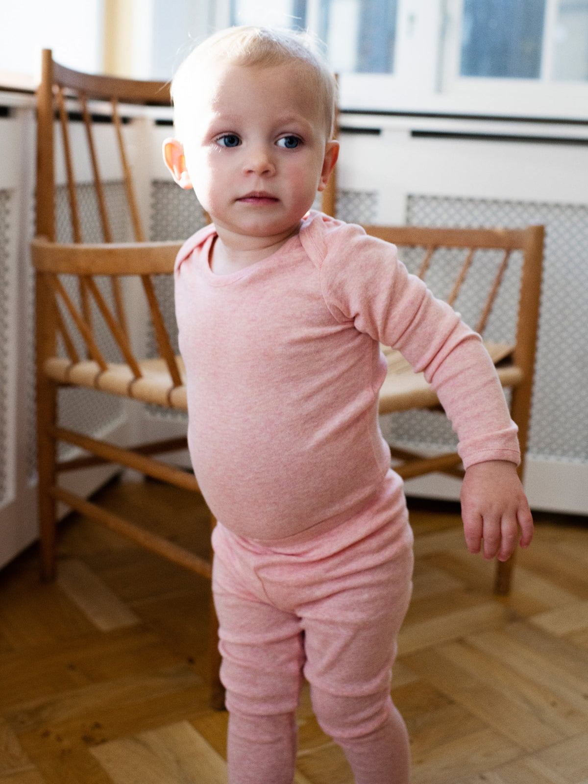 A toddler standing indoors wearing Serendipity's organic cotton coral long-sleeve one-piece bodysuit with matching leggings.