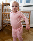 A toddler standing indoors wearing Serendipity's organic cotton coral long-sleeve one-piece bodysuit with matching leggings.