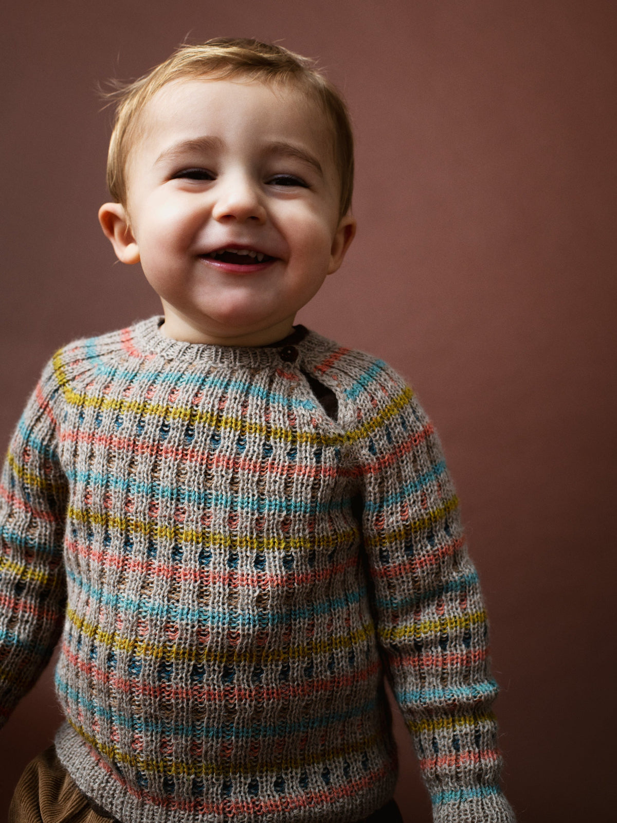 A smiling young child wearing Serendipity Organic's alpaca multicolored rib knit sweater with horizontal stripes of blue, yellow, pink, and brown over a gray base, set against a solid dark brown background.