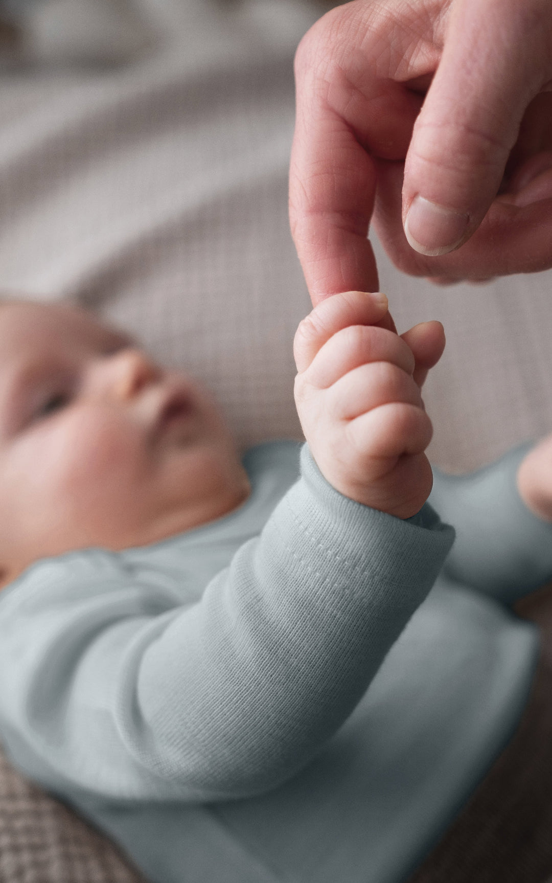 baby in slate blue bodysuit holding adult finger